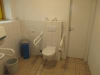 Toilettensitz an einer gekachelten Wand mit einem Hlategriff links und rechts, links in der Ecke des Raumes ein Mülleimer