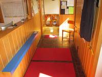 Ein Flur zwischen zwei holzverkleideten Wänden, auf dem Boden liegen rote Schmutzabweiser. Am Flurende ist eine offene Tür