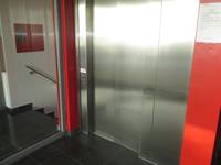 Aufzugtür asu Metall. Zurückversetzt in rot lackierten,breiter Rahmen. Links von Aufzug Glastür mit Blick auf abwärtsfphrende Treppe
