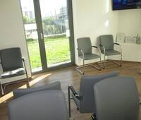 Großer Raum mit Glastür zum Außenbereich. An den Wänden und mittig im Raum stehen grau gepolsterte Stühle.