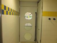  Tür mit drei kreisförmigen Scheiben übereinander angeordnet, an der Tür senkrechte Stange, rechts und links Wände mit gelb-blauen gekachelten Streifen