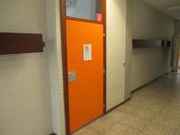 orangefarbige Tür mit braunenmRahmen, darüber Oberlicht, rechts und links von der Tür  weiße Wand mit Garderobenhaken. 
