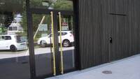 zweiflüglige Glastür in dunklem Metallrahmen und gelben Längsstangen zum Türöffnen. Rects von der Tür ist die dunkelbraune Holzfassade, links eine Glasfront
