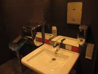 Ein weißes Waschbecken an einer dunklen Wand. Über dem Waschbecken ist ein Spiegel, ein Seifenspender und leicht rechts ein Papierhandtuchspender