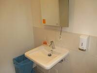 Ein weißes Waschbecken an eienr hellen Wand. Über dem Waschbecken ist ein Spiegel. Rechts davon ist ein Seifenspender an der Wand