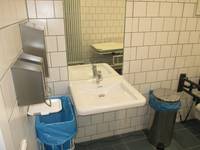 weißes Waschbecken an einer weiß gekachelten Wand, darüber ein Spiegel, rechts davon ein Mülleimer. Links ein Papierhandtuchhalter und zwei Seifenspender,  darunter ein Mülleimer mit einer blauen Mülltüte