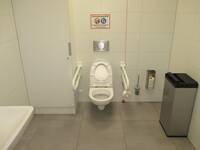 Eine weiße Behindertentoilette mit Handlauf auf jeder Seite. Bei dem Haltegriff auf der rechten Seite steht der Hygienebehälter.