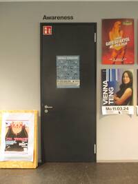 Die Tür und der Türrahmen sind dunkel. In der Türmitte ist ein kleines Plakat angebracht. An der linken oberen Ecke der Tür klebt ein Feuerlöscher-Schild. Rechts an der Wand hängen zwei Poster in vertikaler Anordung. Über der Tür steht an der Wand die schwarze Beschriftung: Awareness.