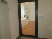 Eine Glastür in einem dunklem Rahmen in einer weißen Wand. Rechts neben der Türe ist eine Klingel/Türöffner