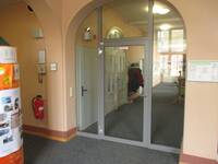 Halbrundes Portal mit Glaswand, mittig darin ist eine Glastür mit einem hellgrauen Rahmen. Dahinter ist ein breiter Flur mit Kleiderhaken und abgehenden Türen.