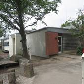 Flachdachgebäude, davor Schulhof mit Bäumen und ein Zugangsweg, am rechten Bildrand eine offene Schranke