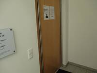 Eine braune Holztür in einer hellen Wand. An der Tür sind 2 DIN A4-Aushänge zu Corona-Maßnahmen