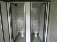 zwei nebeneinander liegende Toilettenkabinen mit offenen Türen, in der Kabine je eine Standtoilette, dahinter Spülkasten. An der Wand WC-Papierrolle