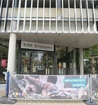 Gebogene Glasschiebetür, darüber hängt ein großes Schild mit der Aufschrift: Zoo Heidelberg.
Vor dem Eingangsbereich stehen Abgrenzungen, die mit Tiermotiven bedruckten Bannern  verkleidet sind