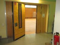 offenstehende Holztür mit eingesetzten Glasscheibe, davor gefliester Boden, dahinter Sporthalle