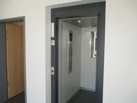 offen stehender Aufzug, weiße Innenkabine. An der Rückwand in der oberen Hälfte angebrachter Spiegel, Bedienelement links.
