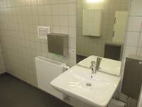 Ein weißes Waschbecken. Darüber ein Spiegel. Links ist eine Heizung und darüber ein Papierhandtuchspender. Rechts davon ist ein Seifenspender