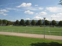 Rasenplatz mit Spielfeldmarkierungen. Am Spielfeldrand ein breiter betonierter Streifen, mit Metallstangen vom Feldabgegrenzt