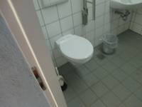 weiße Toilettenschüssel an einer weiß gekachelten Wand, rechtzs davon an der Wand ein Ausschnitt des Haltegriffs und des Waschbecken