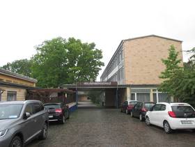 Schulgebäude, mit rechts mehrstöckigem Gebäude, daran links anschließend, ein überdachter Zugang, weiter links ein Teil eines einstöckiges Gebäude, im Vordergrund Zugangsweg mit parkenden Autos rechts und links