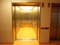 Offener Aufzug mit Taster rechts, der Innenraum ist hell erleuchtet, metallische Wände mit Handlauf, rechts davon ein zweiter geschlossener Auzug mit Taster links 