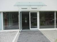 zweiflügelige Glastür mit einem offenen Flügel, davor ein Weg aus Betonverbundsteinen, links und rechts des Weges Parkplätze
