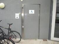 Eine einflügelige Toilettentür, wie die Gebäudewand mit grauen Mosaikfliesen gestaltet. An der linken Seite ist eine senkrecht verlaufende Stange und über die gesamte Türhöhe senkrecht verlaufender Edelstahleinsatz. Auf der Tür ist ein großes Rollstuhlsymbol auf einem weißen Schild. Links neben der Tür ist ein kleines Symbol für einen Wickeltisch.