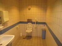 Eine weiße Toilette in einem weiß gekacheltem Raum. In dem Raum ist auf halber Höhe ein blauer Streifen der ringsrum läuft.