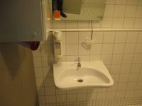 Ein weißes Waschbecken an einer weiß gekachelten Wand. Darüber ein Spiegel, ein Seifenspender und links davon ein Handtuchhalter