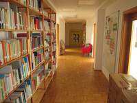 Ein gerade verlaufender Flur, mit Bücherregalen auf der linken Seite und Türen auf beiden Seiten. Im Hintergrund rechts steht im Flur ein rotes Sofa.
