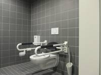 Hänge-WC mit Handläufen rechts und linksan einer dunkel gekachelten Wand, ausserdem eine Toilettenbürste und eine Drucktastte an der Rückwand                