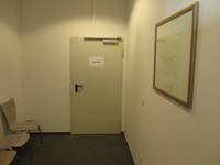 hellgrau Metalltür mit einer schwarzen Klinke, rechts eine Wand mit einem Bild, links stehen an der Wand 3 Stühle