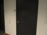 Eine dunkle Türe in einer hellen Wand. Rechts von der Türe ist ein kleines Rollstuhl-Symbol an der Wand
