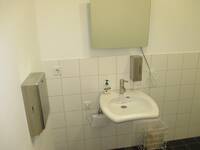 Weißes Waschbecken an einer weiß gekachelten Wand, Über dem Waschbecken hängt ein Spiegel, an der Wand links daneben hängt ein Papierhandtuchspender