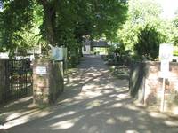 Ein offenstehendes Tor mit je einem Pfeiler rechts und links. Nach dem Tor führt ein Weg geradeaus bis zu einer Kapelle im Hintergrund. Rechts und links sind Bäume