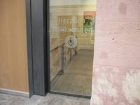 Glastür mit Rollstuhlfahrersymbol in der Mitte. Der Eingang ist etwas zurückgesetzt. Rechts von der Tür beginnt die Sandsteinfassade des Gebäudes