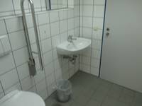 Waschbecken an einer gekachelten Wand links davon ein Haltegriff und die Toilette, rechts davon eine geschlossene Tür