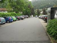  Parkplatz mitparkenden Autos rechts und links. Vorne rechts ist eine Behindertentoilette. Bei den Parkplätzen sind Büsche und Bäume 