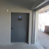Tür mit Toilettensymbolen  umgeben von grauer Wand, rechts davon offen stehendes Tor