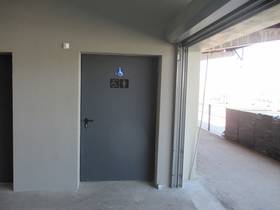 Tür mit Toilettensymbolen  umgeben von grauer Wand, rechts davon offen stehendes Tor