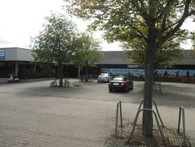 Großer L-Förmiger Fachdachbau, davor ein Parkplatz und etliche Bäume