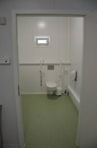 weiße Hängetoilette an weiß gekachelter Wand. Links und rechts 2 hochgeklappte Haltegriffe. Über der Toilette ist ein kleines Oberlicht.
