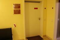 Weiße Eingangstür mit Rundknopf, die Wand ist gelblich und wenig kontrastreich zur Türe, links ist ein elektronischer Türöffner und über dem Türöffner ist das Praxisschild, links ist ausserdem noch ein Sessel