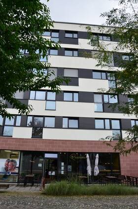 Sechsstöckiger großer Neubau mit Wohnungen ab dem 1. OG.  Fassade im Erdgeschoss mit rotem Sandstein verkleidet. Glasfront mit Eingnagstür zu Restaurant, Tische und Stühle im Außenbereich