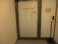 Eine Glastür, innen weiß beklebt, mit einem dunklem Metallrahmen. Auf der Tür ist ein roter Schriftzug: Orthopädische Praxis. Rechts neben der Tür steht ein Spender für Desinfektionsmittel.