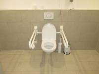 an einer gekachelten Wand eine weiße Toilette mit Haltegriffen rechts und links, rechts daneben ein Mülleimer und eine Toilettenbürste