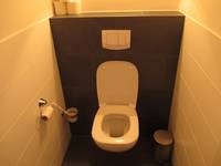 weiße Toilette an schwarzer Wand, rechts und links sind die Wände weiß gekachelt. An der linken Wand ist ein WC-Papierrollenhalter und WC-Bürste, in der rechten Ecke steht ein Mülleimer