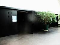 Im Zentrum aif der linken Seite ist der Eingansbereich zum Saal mit mehreren schwarzen Türen in einer schwarzen Front. Eine Tür steht offen, rechts daneben steht eine große Grünpflanze.