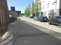 ein breiter Weg, links davon die Holzverschalung eines Gebäudes, rechts sind Parkplätze mit parkenden Autos. Neben den Parkplätzen steht ein mehrstöckiges Wohnhaus