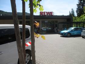 Im Vordergrund ist ein Parkplatz auf dem ein einstöckiges Gebäude steht mit einem großen Schriftzug REWE